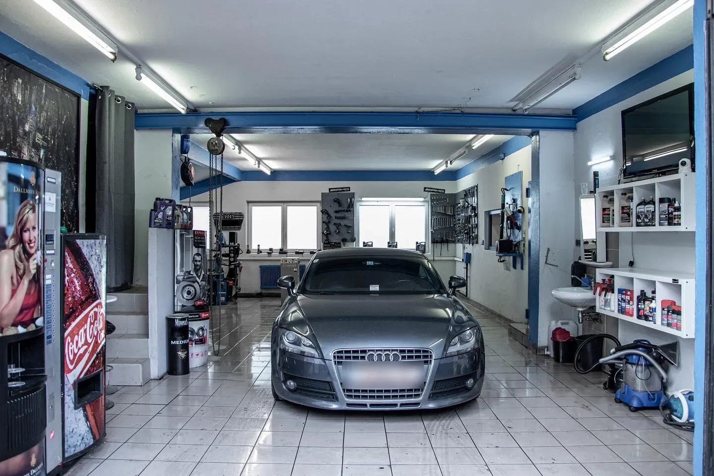 Blick in die helle Werkstatthalle von Kingdom of Cars: ein grauer Audi TT steht frontal auf dem gefliesten Boden, links und rechts Werkzeugwände und Regale